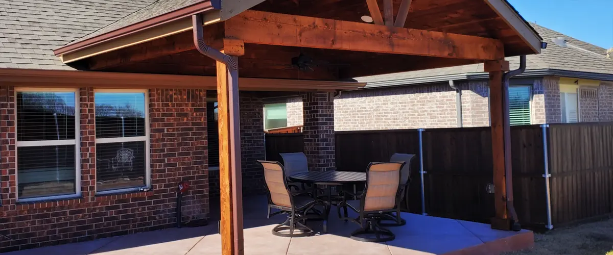 Covered patio with stained wood pergola, ceiling fan, and outdoor dining set on stamped concrete in brick backyard