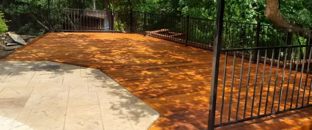 Custom wood deck with black metal railing in a wooded backyard next to a stone patio and pool area