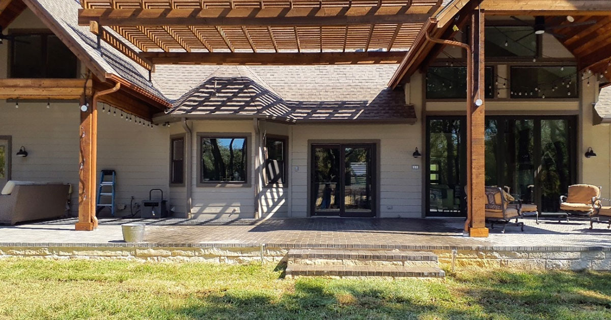 Large backyard patio with wood pergola, brick pavers, and outdoor lounge seating attached to modern farmhouse-style home