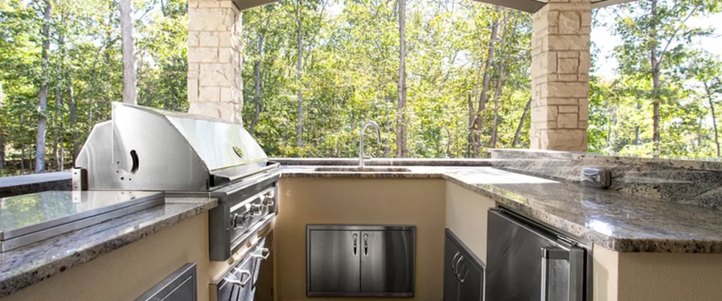 outdoor kitchen with stainless steel grill granite countertops stone pillars wooded backdrop