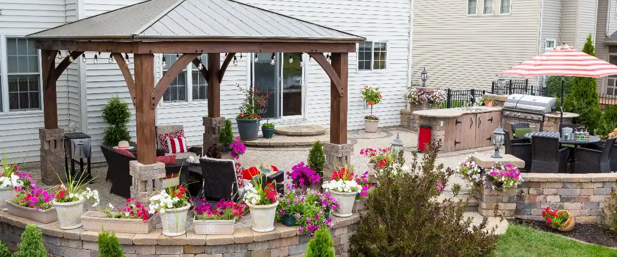 A covered patio in a backyard surrounded by colorful flowers and a grill nearby, creating a cozy outdoor space.