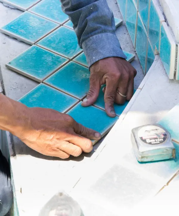 A man is carefully laying tiles on a pool floor, focused on his work with tools nearby