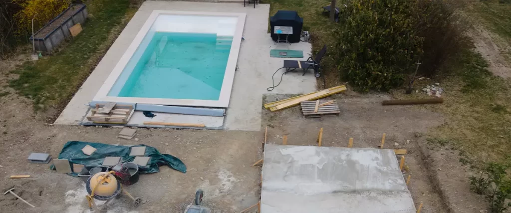 An overhead view of an inground pool installation project showing the finished pool shell on the left, an excavated area and freshly poured concrete slab on the right, and construction materials and equipment nearby.