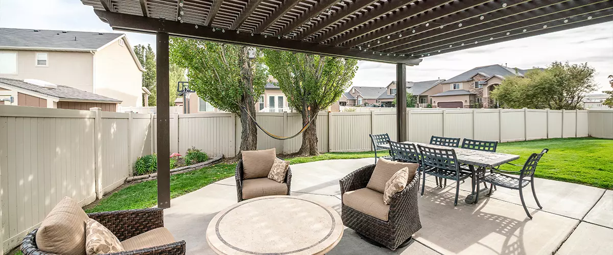 A spacious backyard patio with a dark louvered pergola covering dedicated dining and seating areas. The patio features a long dining table with chairs, a round fire pit table with cushioned armchairs, and a woven hammock strung between two trees, all within a fenced yard with green grass and surrounding houses