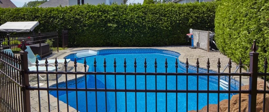 An outdoor swimming pool area securely surrounded by a black wrought iron fence with decorative finials, set against a backdrop of a tall, dense green hedge.