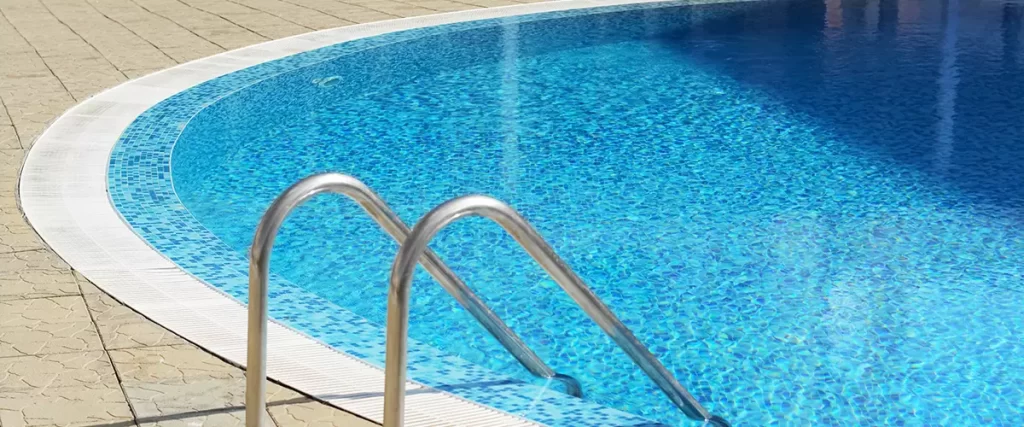 A close-up view of a curved, blue tiled swimming pool edge with stainless steel handrails of a ladder leading into the water, surrounded by light beige textured paving stones.