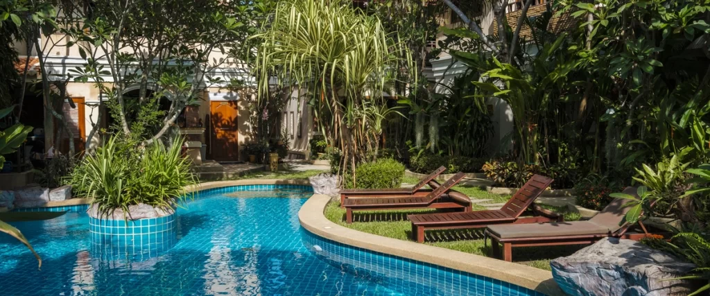A winding swimming pool with blue mosaic tile interior, surrounded by dense, lush tropical landscaping, grass, and two dark wooden lounge chairs.