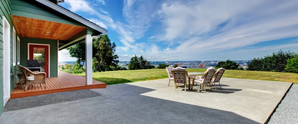 A large, simple concrete patio extending from a house with a wooden porch, offering expansive views of a distant city skyline and open landscape.