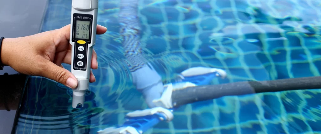A close-up of a hand holding a digital salt meter to test the water quality and salinity in a tiled swimming pool. The meter displays a reading of 3.160.