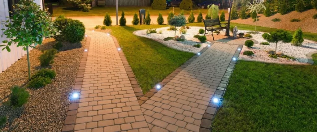 Two parallel interlocking brick paver walkways, lit by small embedded ground lights at night, separating a lawn and a decorative garden area.
