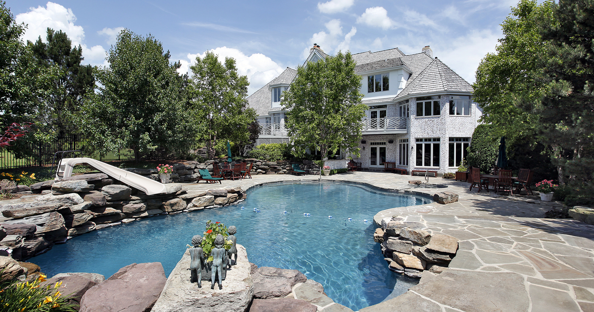 A sprawling luxury home backyard featuring a lagoon-style inground pool with a rock waterfall, a small slide, flagstone patio, and lush landscaping, illustrating current pool design trends.