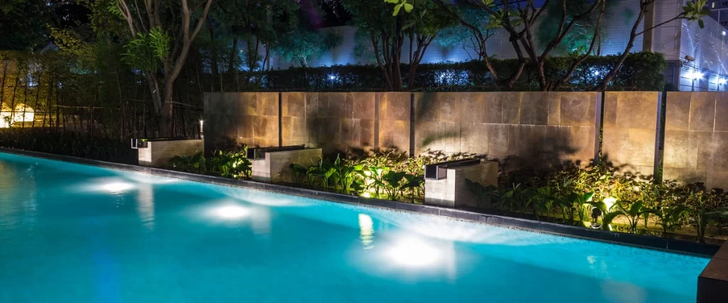 A beautifully lit, modern, rectangular swimming pool at night, featuring submerged lights and surrounded by a sleek privacy wall and lush planters.