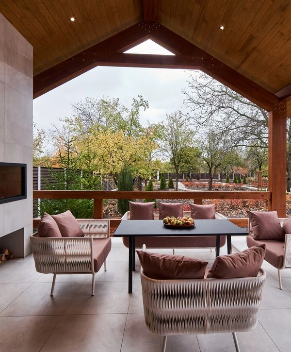 A stylish modern covered patio features a high wooden ceiling, an outdoor fireplace, comfortable woven outdoor furniture with rose-colored cushions around a black dining table, and offers a panoramic view of a landscaped backyard with trees and a fence