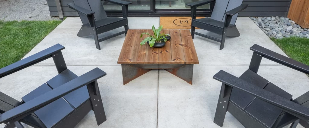 A modern, simple patio made of large-square light gray stamped concrete, furnished with four black Adirondack chairs and a rustic wooden coffee table.