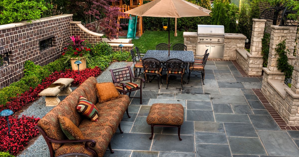 Luxurious backyard patio featuring dark slate pavers, a built-in stone grill station, and an outdoor living area with a sofa, chairs, and dining set under an umbrella. This showcases a high-end paver patio design.