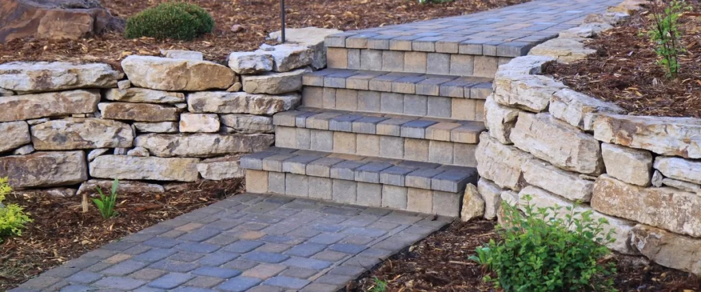 A curved paver walkway leading up to a set of stairs made of multicolored pavers, framed by natural stacked stone retaining walls in a landscaped garden bed.
