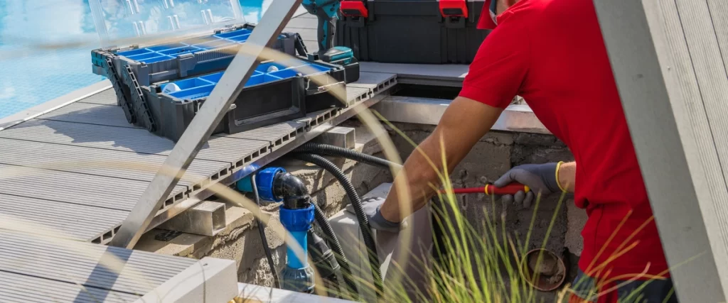 A pool maintenance worker, wearing a red shirt and gloves, examining and adjusting the plumbing and pipes in an underground vault beside a deck.