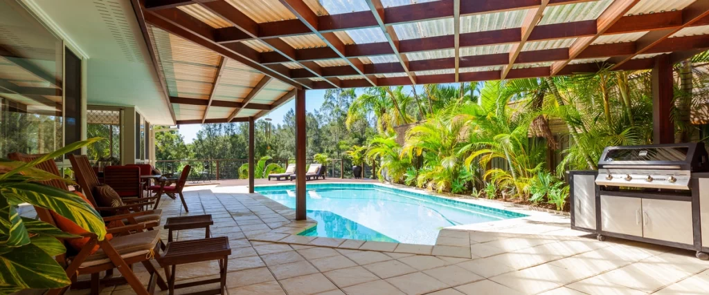 A rectangular swimming pool bordered by a light-tiled patio, featuring a covered pergola area with wood chairs, and a stainless steel outdoor kitchen/grill setup.