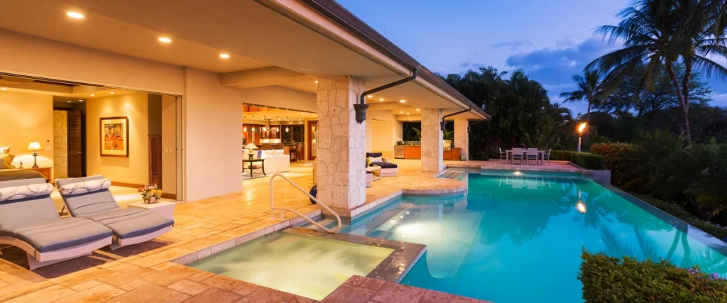 A luxurious residential backyard pool and integrated hot tub area at twilight, surrounded by stone paving and featuring warm interior and exterior lighting.