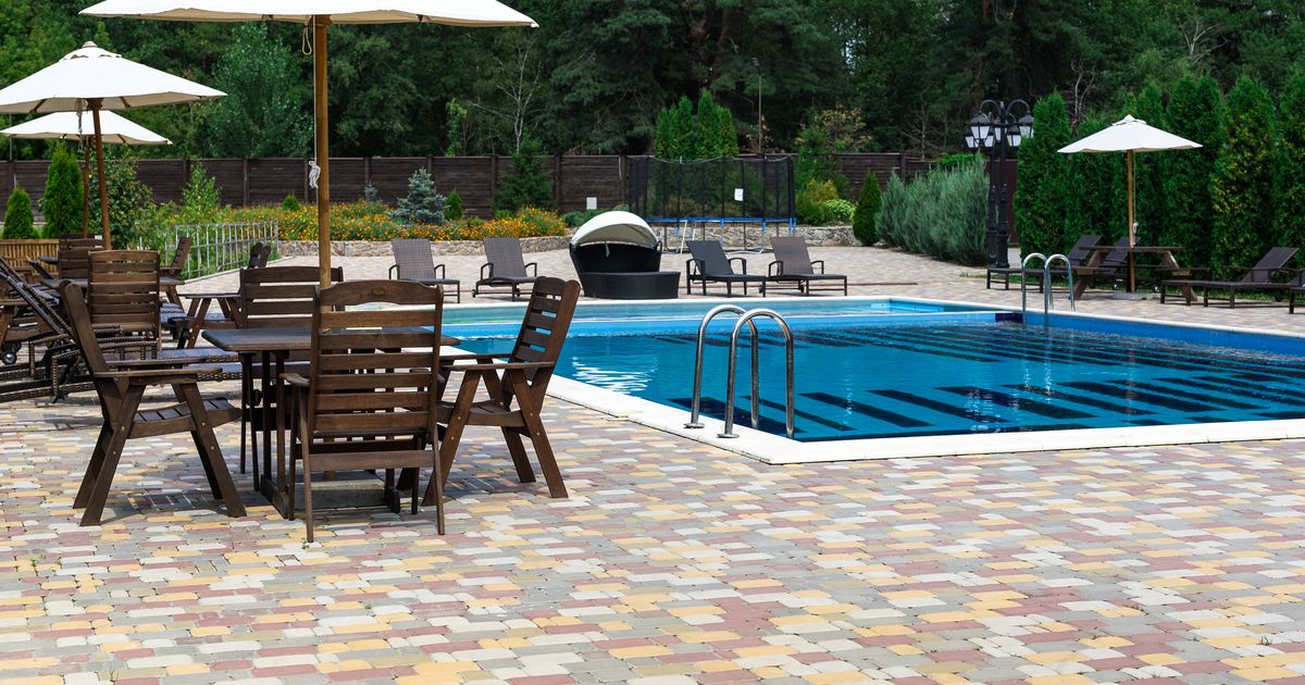 An inviting outdoor resort or community swimming pool with prominent metal safety ladders, surrounded by a colorful paved patio, lounge chairs, and a visible perimeter fence in the background, showcasing essential pool safety features.