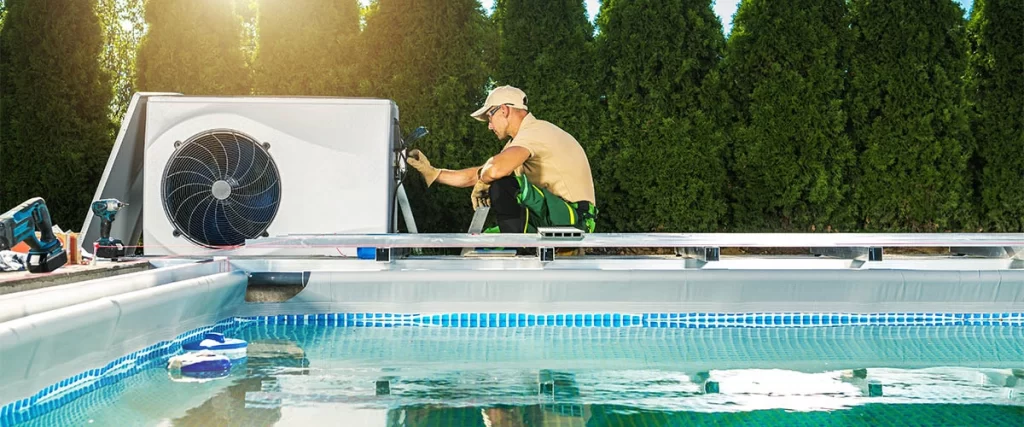 A technician installing or servicing a large, modern white outdoor pool heat pump system next to an in-ground swimming pool on a sunny day.