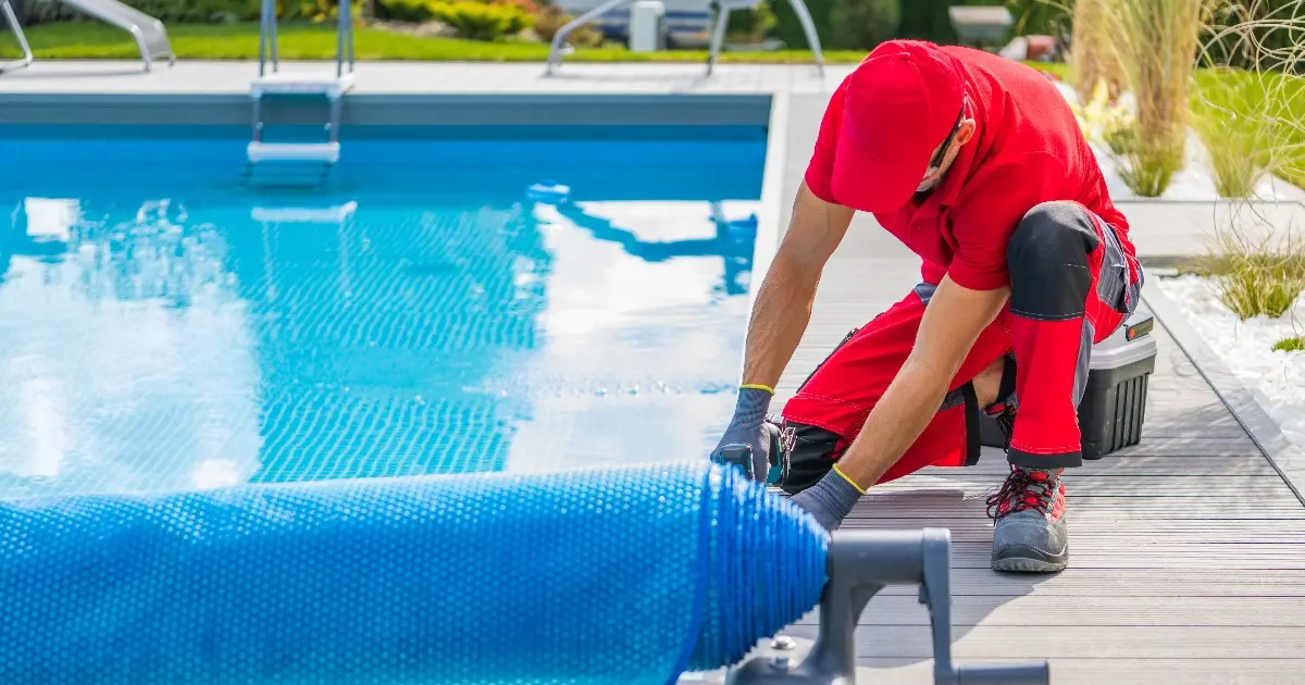 a pool service technician covering an in-ground swimming pool with a blue bubble-style solar cover using a roller system