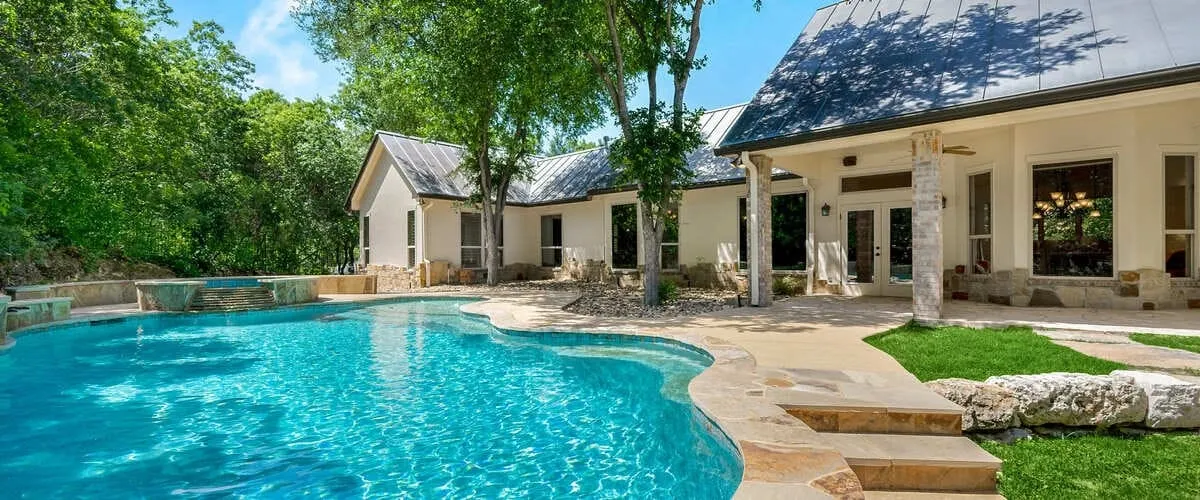 Large custom freeform lagoon-style inground swimming pool with natural stone coping, a raised hot tub, and a spacious tan stone patio surrounding a white ranch-style home.