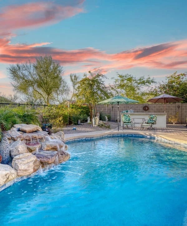 A vibrant backyard swimming pool at sunset featuring a cascading natural stone waterfall, patio umbrellas, and a poolside bar area under a colorful sky.