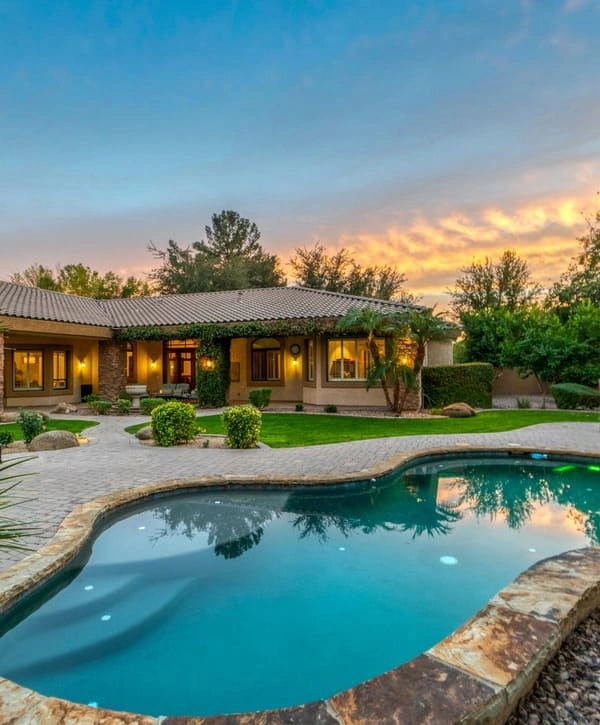A luxury custom-shaped inground swimming pool with natural stone coping and integrated steps, set against a Mediterranean-style villa during a golden hour sunset with lush green landscaping.
