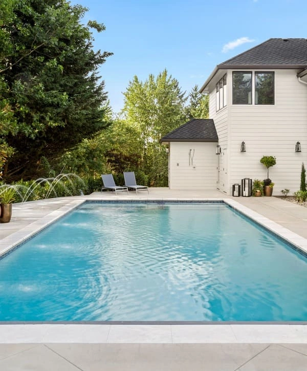 A luxury modern rectangular backyard swimming pool featuring integrated water fountains, a light stone deck with lounge chairs, and a white farmhouse-style home in the background.