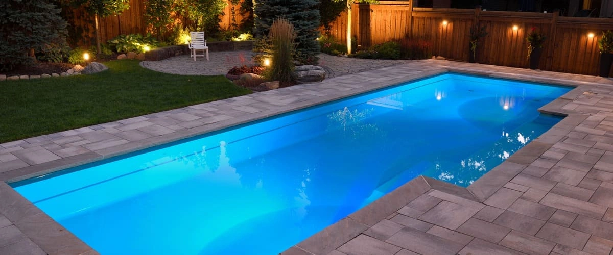 A sleek rectangular swimming pool illuminated by blue internal LED lights at night, surrounded by grey stone patio pavers and a dark wood privacy fence with ambient garden lighting.