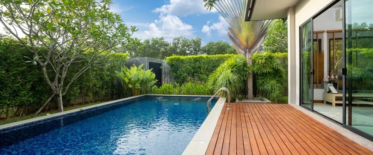 A private residential lap pool bordered by a rich wooden deck and surrounded by dense tropical greenery, palm trees, and manicured hedges.