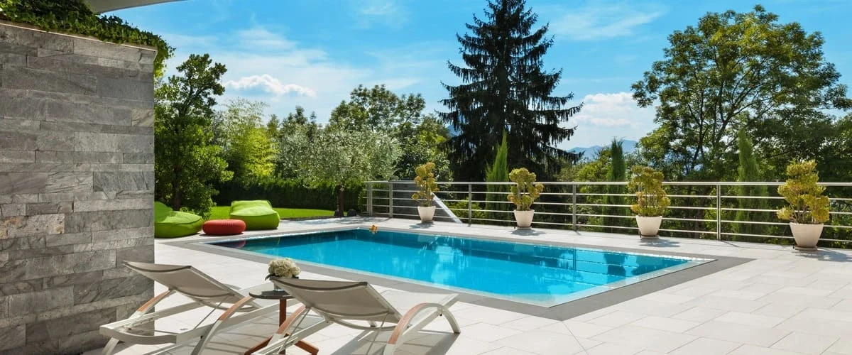 A bright elevated terrace pool featuring white stone tiling, modern white lounge chairs, and a metal safety railing overlooking a dense green forest under a clear blue sky.
