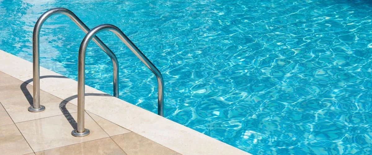 Close-up of a double-handle stainless steel swimming pool entry ladder installed on a light-colored travertine tile deck with clear shimmering blue water.