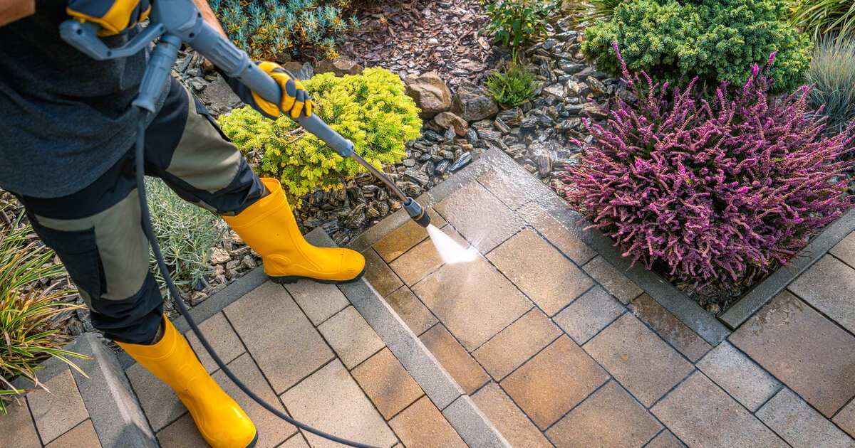 A person wearing yellow boots and protective gear demonstrating the best way to clean patio pavers using a professional pressure washer on a backyard walkway.