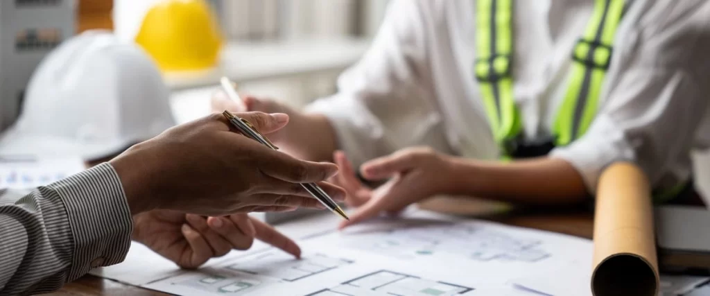 Two professionals in a bright office pointing at sections of a large paper blueprint during a consultation, with safety helmets visible in the blurred background.