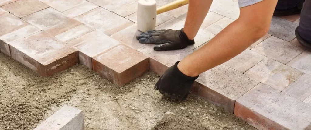 A contractor wearing black gloves using a rubber mallet to carefully level brown stone pavers during a patio installation.