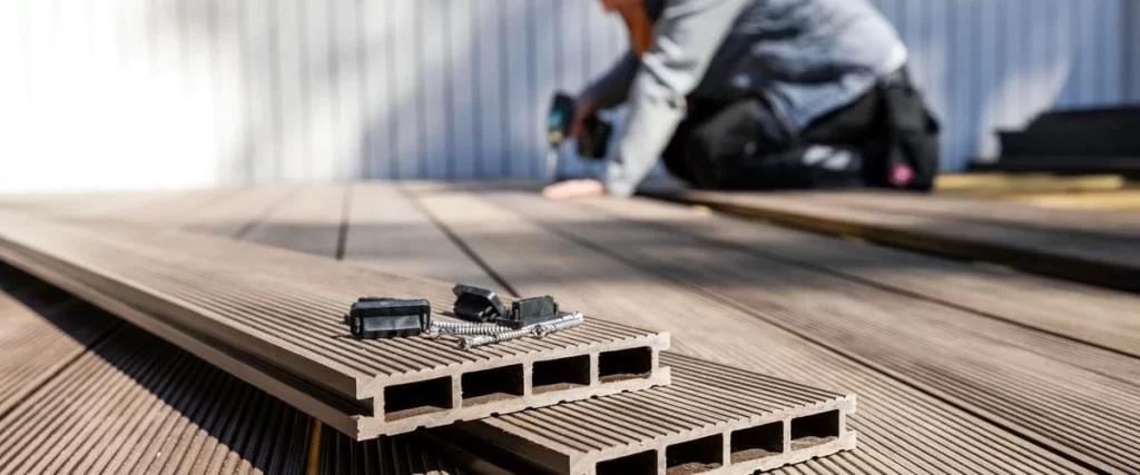 A close-up view of brown hollow-core composite decking boards featuring hidden fasteners and stainless steel screws on a terrace being installed by a professional contractor using a power drill.