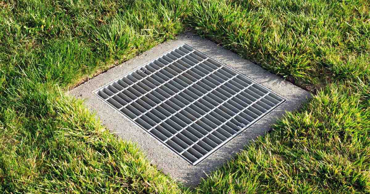 A high-angle shot of a square galvanized steel grate installed in a concrete base, demonstrating efficient patio drainage options for managing water runoff in a green grass lawn.