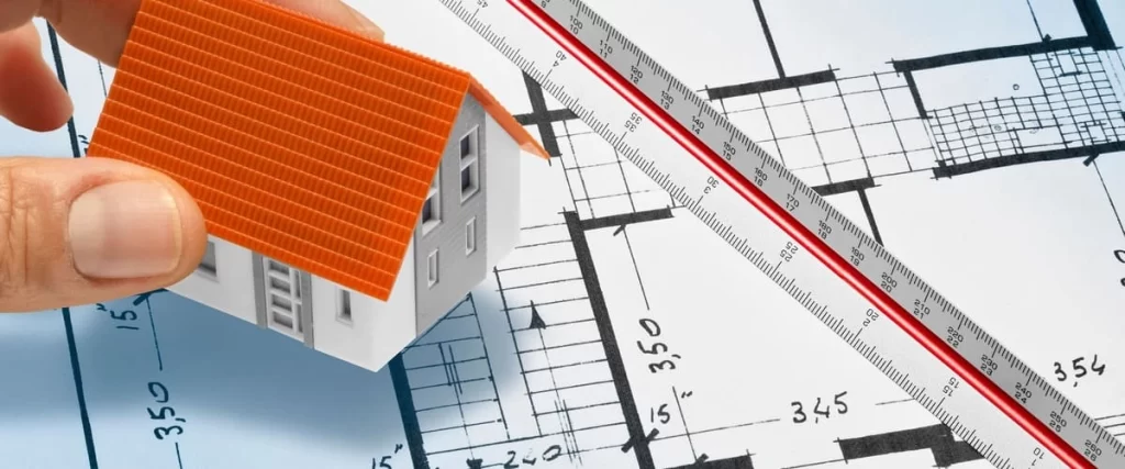 A close-up view of a hand holding a small 3D model of a white house with an orange roof, positioning it over a technical architectural drawing next to a scale ruler.