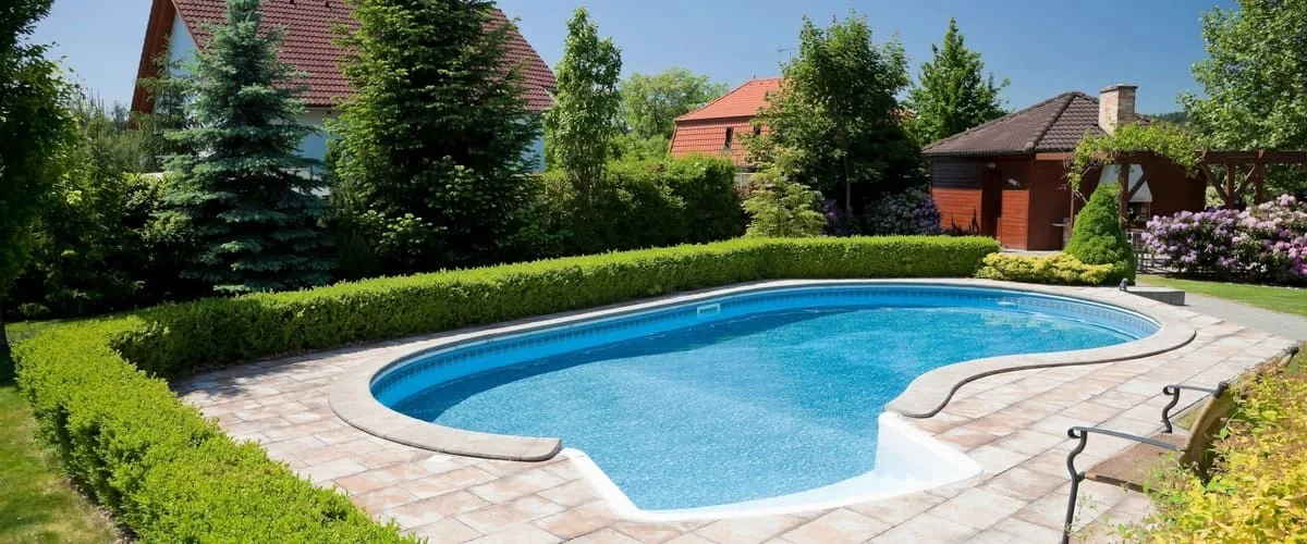 A kidney-shaped blue swimming pool surrounded by light-colored stone tiling and a neatly trimmed low green hedge in a residential backyard.