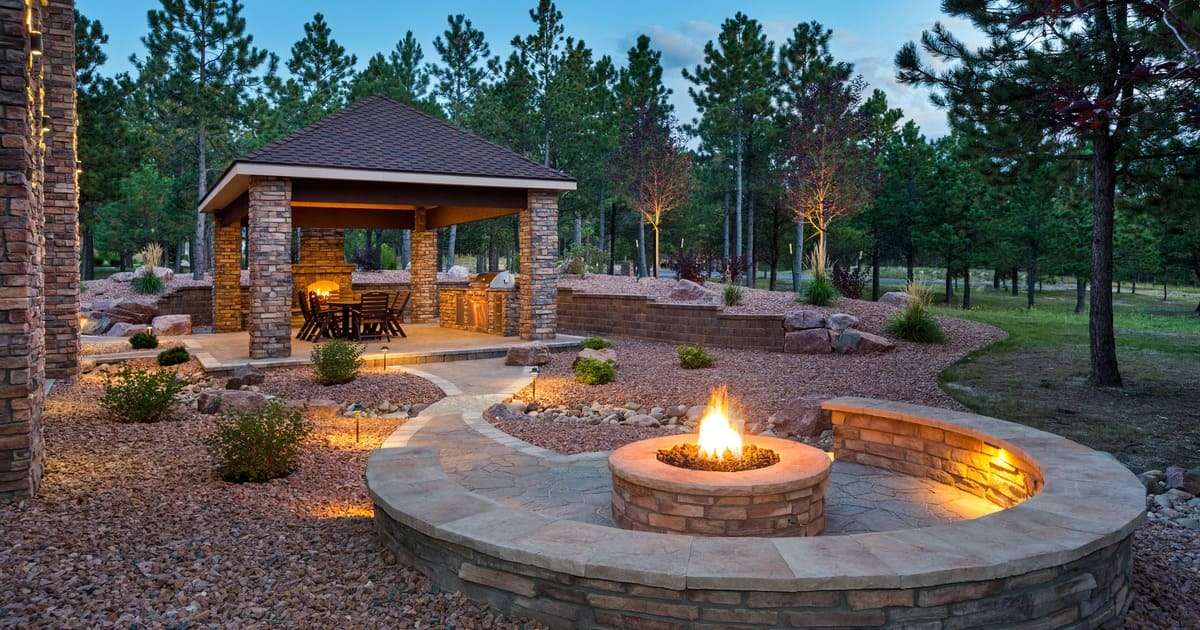 A beautifully landscaped outdoor living area at dusk featuring stone masonry, a circular built-in seating area around a glowing fire pit, and a covered pavilion with an outdoor kitchen, perfect for backyard patio ideas with fire pit inspiration.