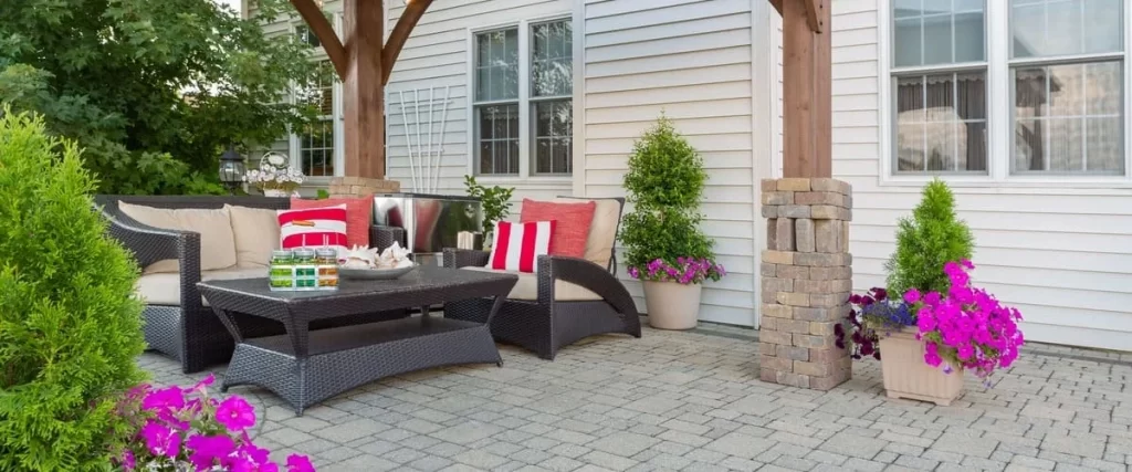 A cozy outdoor seating area under a pergola with black wicker sofas, pink flowers, and clean stone paver flooring.