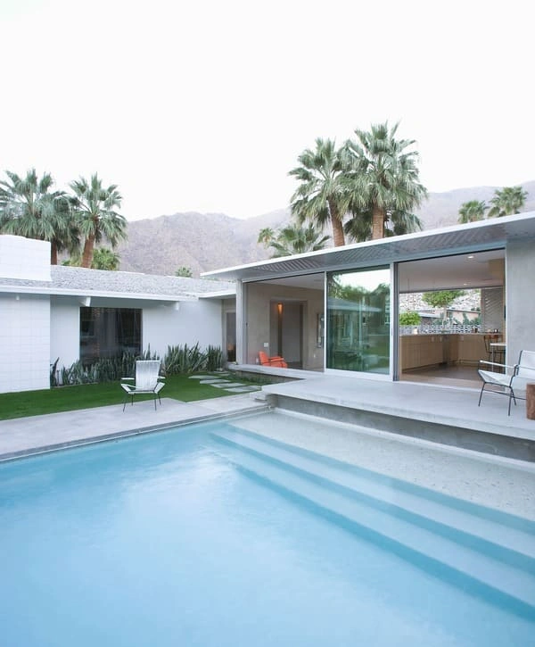 An architectural white house with expansive glass walls and an open living area reflecting in the calm water of a large swimming pool with wide steps.