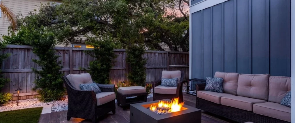 A modern outdoor living space at twilight featuring a grey sectional sofa with patterned pillows and a square concrete fire pit with blue glass media on a wood-look tile deck.