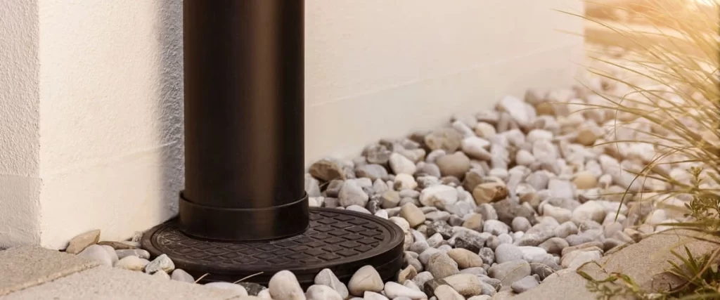 A close-up view of a vertical black PVC pipe for french drains connected to a round drainage grate, surrounded by small grey river stones and decorative pebbles next to a white exterior wall and stone patio pavers.
