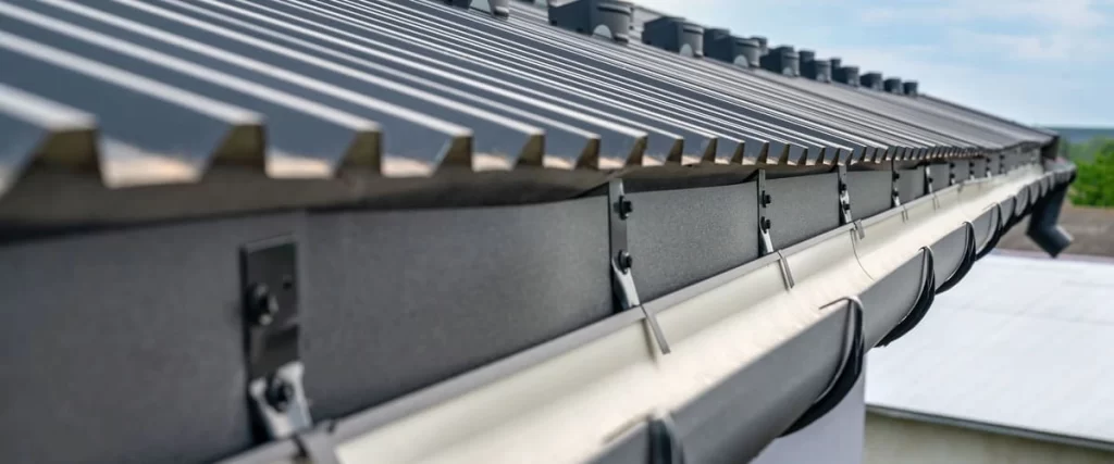 A close-up view of a professional installation of white seamless rain gutters attached to a dark charcoal grey corrugated metal roof with secure mounting brackets.