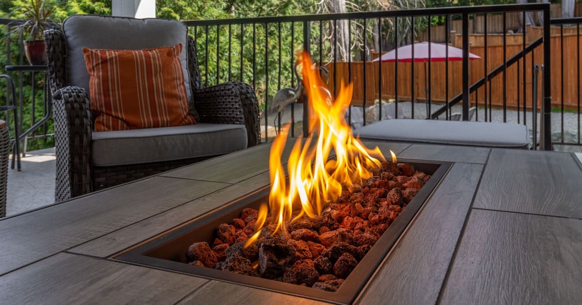 A rectangular propane fire pit table with lava rocks burning on a covered patio with comfortable wicker chairs and a striped orange pillow.