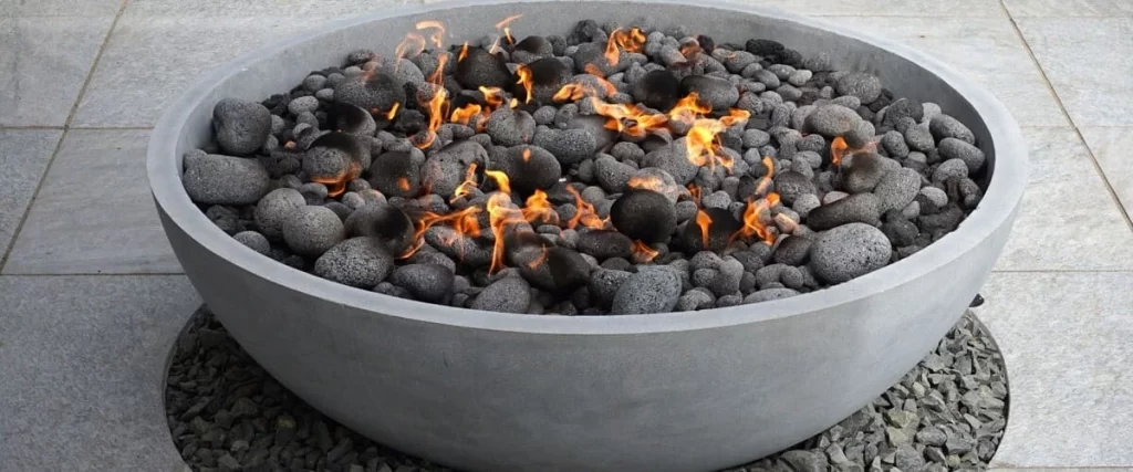 A modern round grey concrete fire pit sits on a stone patio filled with black lava rocks and small flickering orange flames.