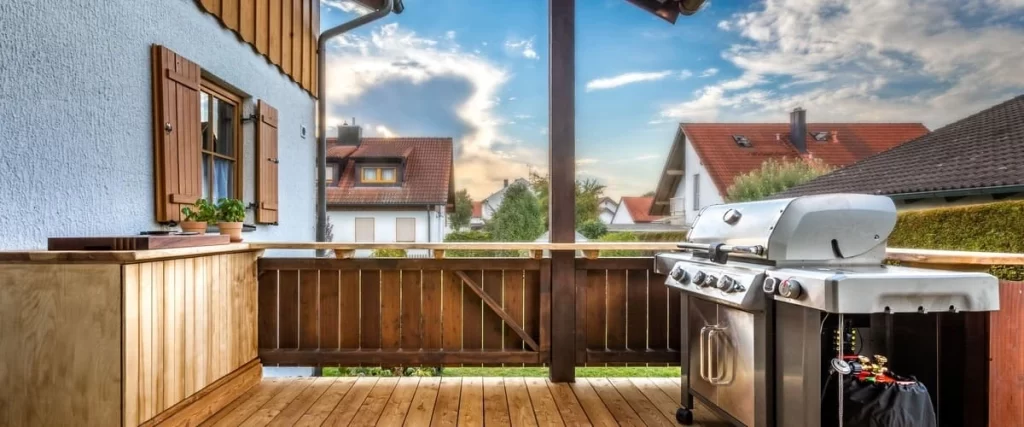 A functional wooden deck setup for cooking, featuring a large stainless steel gas grill and a custom wooden outdoor kitchen counter against a rustic privacy fence.